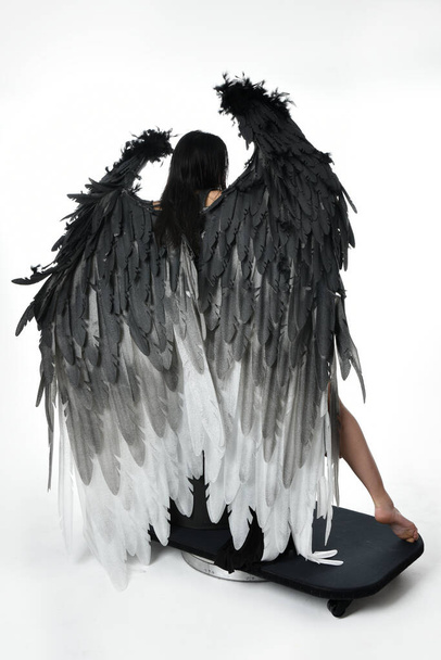 full length portrait of beautiful asian model with dark hair, wearing black gothic skirt costume, angel feather wings with horned headdress. Sitting pose  isolated on studio background. - Photo, image