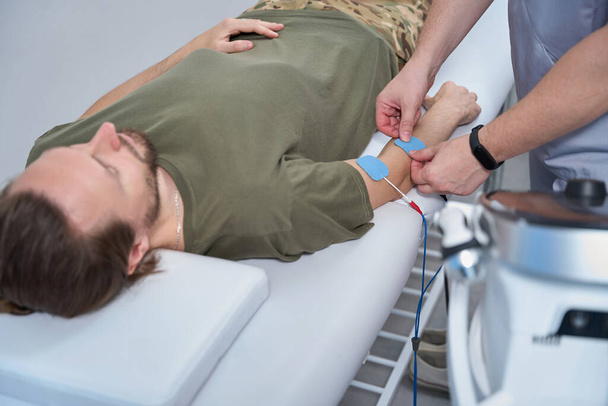 Military man lies on a couch on a hardware procedure, a physiotherapist attaches electrodes to the patient arm - Foto, immagini