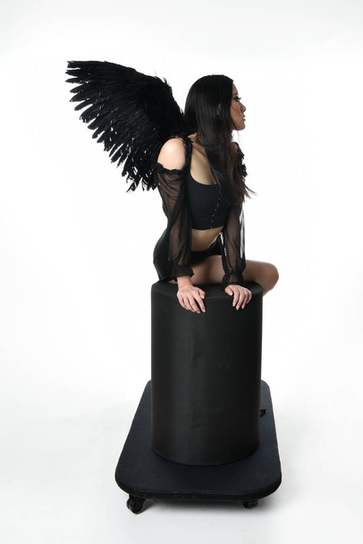 full length portrait of beautiful asian model with dark hair, wearing black gothic skirt costume, angel feather wings with horned headdress. Sitting pose  isolated on studio background. - Foto, afbeelding
