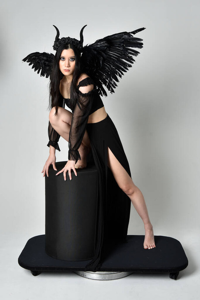 full length portrait of beautiful asian model with dark hair, wearing black gothic skirt costume, angel feather wings with horned headdress. Sitting pose  isolated on studio background. - Foto, Bild