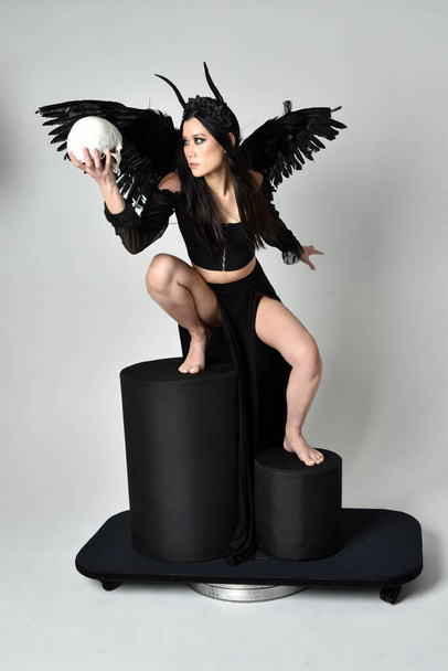 full length portrait of beautiful asian model with dark hair, wearing black gothic skirt costume, angel feather wings with horned headdress. Sitting pose  isolated on studio background. - Foto, Imagen