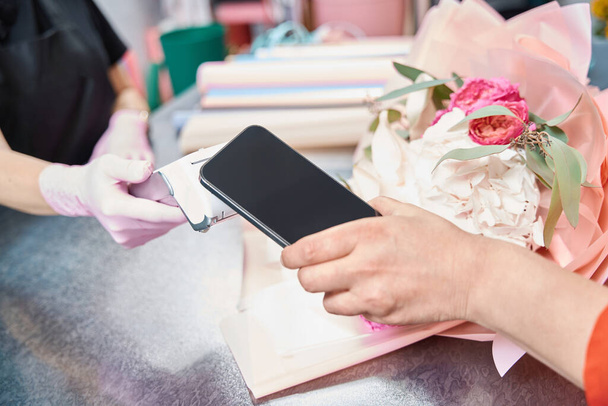 Woman hand holds phone near terminal to pay for ordered bouquet in flower boutique. Nearby lies ready-made bouquet of flowers. - Photo, image