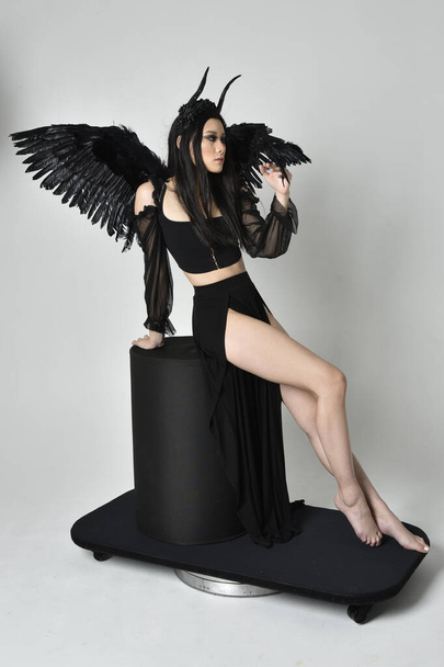 full length portrait of beautiful asian model with dark hair, wearing black gothic skirt costume, angel feather wings with horned headdress. Sitting pose  isolated on studio background. - Фото, изображение
