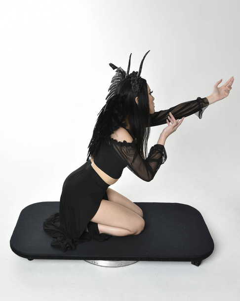 full length portrait of beautiful asian model with dark hair, wearing black gothic skirt costume, angel feather wings with horned headdress. kneeling  pose  isolated on studio background. - Foto, Imagem