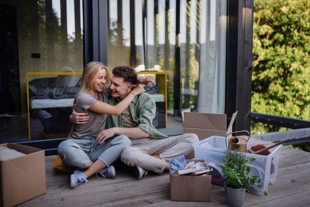 A cheerful young couple moving in their new tiny house in woods sitting on floor on terrace. Conception of moving and sustainable living. - Photo, Image