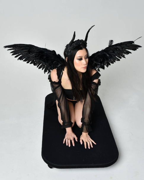 full length portrait of beautiful asian model with dark hair, wearing black gothic skirt costume, angel feather wings with horned headdress. kneeling  pose  isolated on studio background. - Фото, зображення