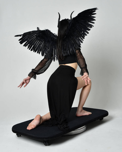 full length portrait of beautiful asian model with dark hair, wearing black gothic skirt costume, angel feather wings with horned headdress. kneeling  pose  isolated on studio background. - Fotoğraf, Görsel