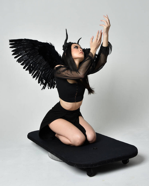 ολόσωμο πορτρέτο του όμορφου ασιατικού μοντέλου με σκούρα μαλλιά, φορώντας μαύρη γοτθική φούστα κοστούμι, φτερά αγγέλου φτερό με κέρατο κεφαλόδεσμο. γονατιστή στάση απομονωμένη στο φόντο στούντιο. - Φωτογραφία, εικόνα