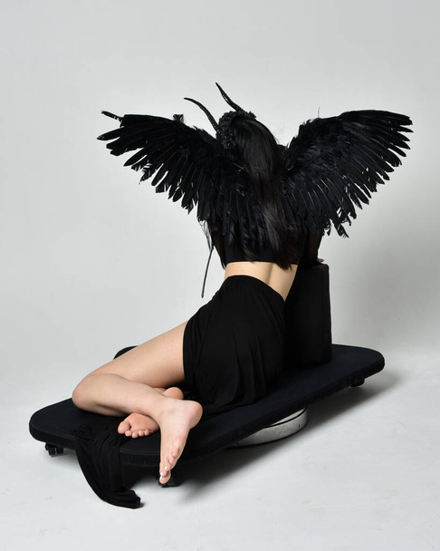 full length portrait of beautiful asian model with dark hair, wearing black gothic skirt costume, angel feather wings with horned headdress. kneeling  pose  isolated on studio background. - Photo, image