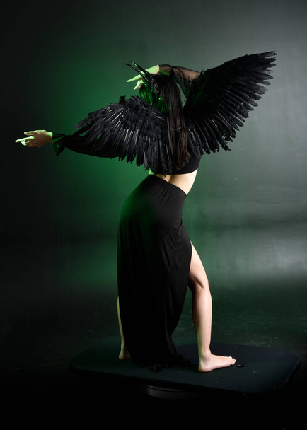 portrait of beautiful asian model with dark hair, wearing black gothic skirt costume, angel feather wings with horned headdress. Posing with gestural hands  on dark silhouette  studio background. - Foto, Imagem
