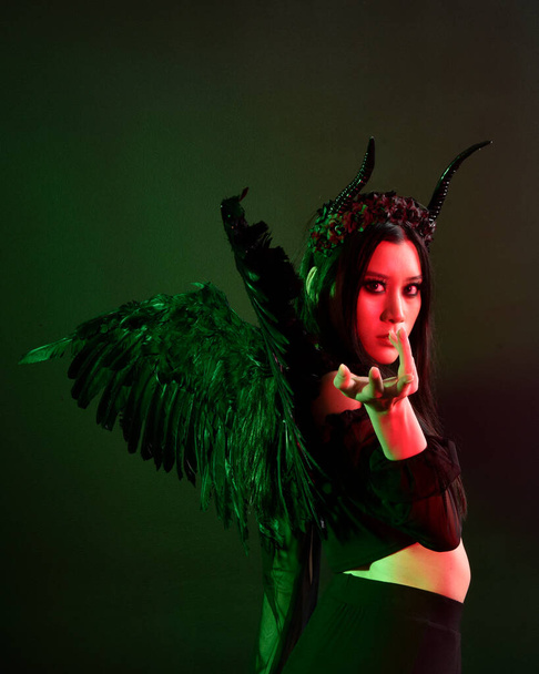 portrait of beautiful asian model with dark hair, wearing black gothic skirt costume, angel feather wings with horned headdress. Posing with gestural hands  on dark silhouette  studio background. - 写真・画像