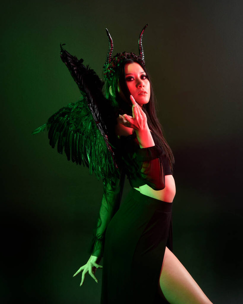 portrait of beautiful asian model with dark hair, wearing black gothic skirt costume, angel feather wings with horned headdress. Posing with gestural hands  on dark silhouette  studio background. - Foto, immagini