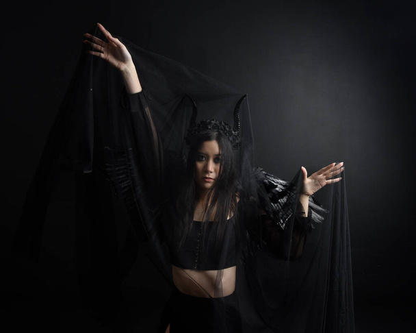 portrait of beautiful asian model with dark hair, wearing black gothic skirt costume, angel feather wings with horned headdress. Posing with gestural hands  on dark silhouette  studio background. - Photo, image