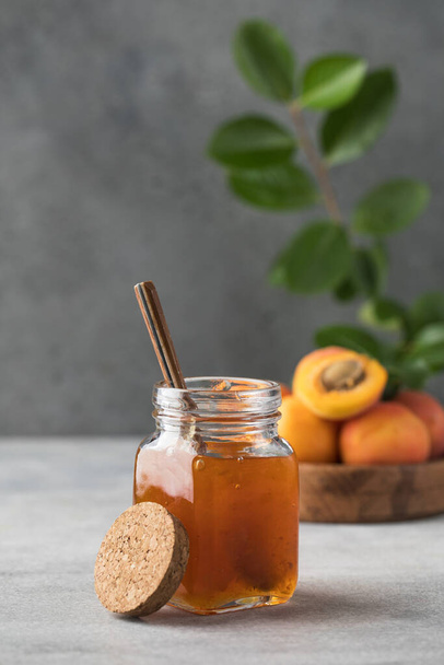 A jar with apricot jam and a spoon close-up on a gray background. In the background is a plate with fresh apricots and a branch with green leaves. Fruit preservation. - Fotó, kép