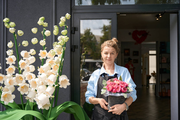 Portrait of cute female florist in apron with bouquet of peony roses in box in front of flower shop - Фото, изображение