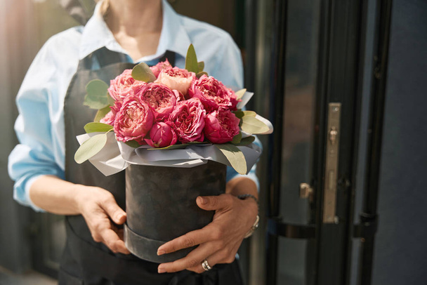 Cropped photo of woman holding luxury bouquet of peony roses in black box in front of flower boutique - Фото, изображение