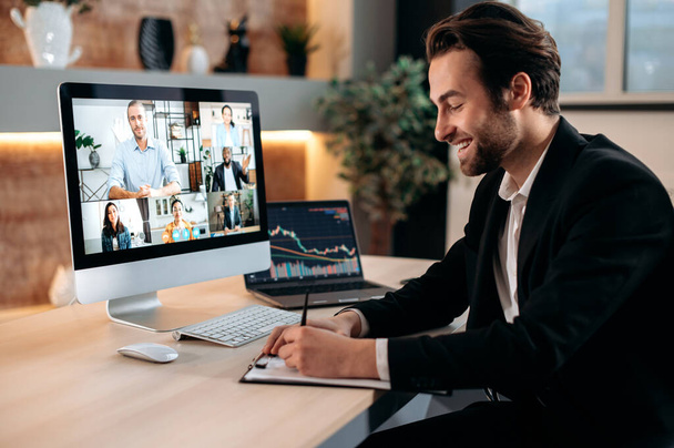 Successful positive Caucasian business man in suit, sit at a workplace, negotiating with partners by video call , discussing new project, terms of cooperation, takes notes. Video conference in office. - Фото, зображення