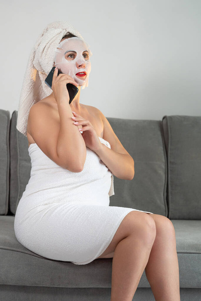 A woman is talking on the phone at home, a woman with a mask on her face and a towel is sitting on the sofa, photo for advertising. - Foto, imagen