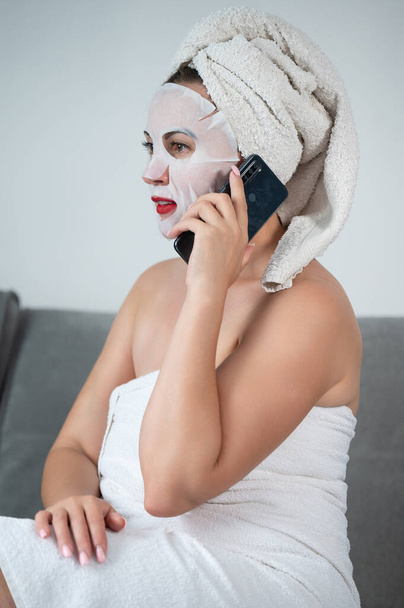 A woman is talking on the phone at home, a woman with a mask on her face and a towel is sitting on the sofa, photo for advertising. - 写真・画像
