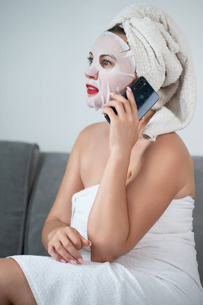A woman is talking on the phone at home, a woman with a mask on her face and a towel is sitting on the sofa, photo for advertising. - Fotografie, Obrázek