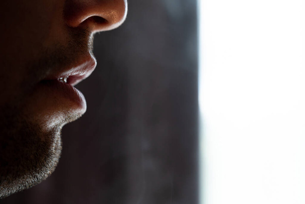 Соблазнительные мужские губы, выдувающие сигаретный дым, легкая щетина на бороде, портрет мужских губ и дым на черном фоне. - Фото, изображение
