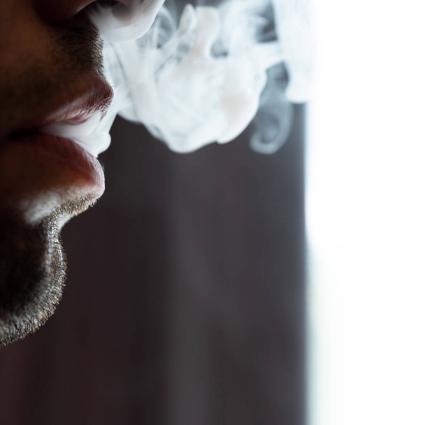 Seductive male lips blowing cigarette smoke, light stubble on beard, portrait of male lips and smoke on black background. - Фото, изображение