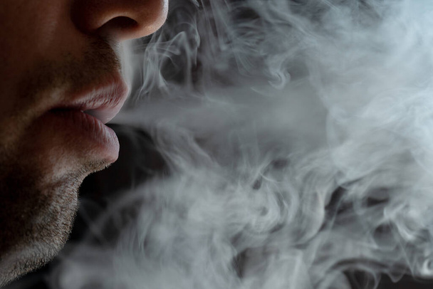 Γοητευτικά αρσενικά χείλη που φυσούν καπνό τσιγάρου, ελαφρά γένια, πορτραίτα αρσενικών χειλιών και καπνός σε μαύρο φόντο. - Φωτογραφία, εικόνα
