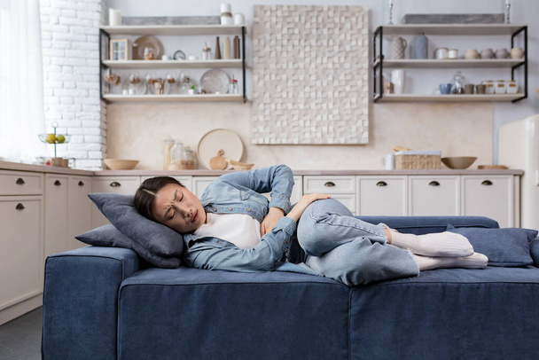 Sick and sad woman lying on the sofa at home in the living room, Asian woman has severe stomach pain, holding her side with her hands - Foto, immagini
