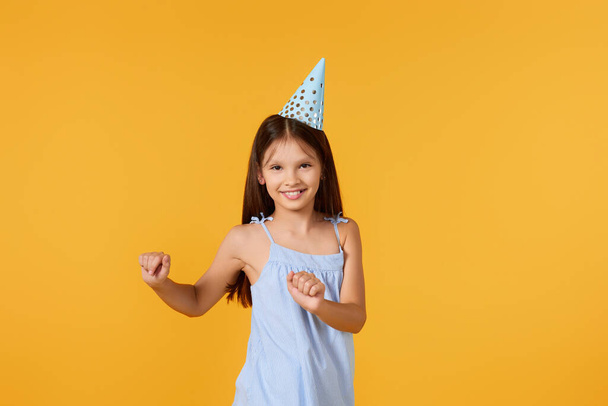 happy birthday child girl in blue dress and party hat dancing on yellow background - Photo, Image