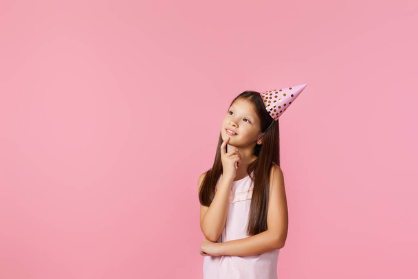 cute little girl with party cone making wish and think about something on her birthday on pink background. copy space - Fotografie, Obrázek