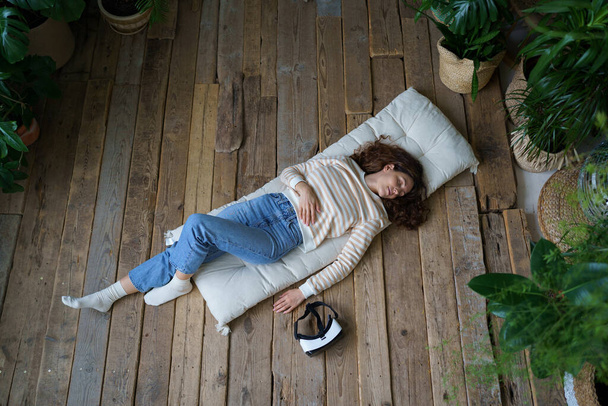 Young peaceful woman sleeping on wooden floor, resting after using VR helmet, female fell asleep while using virtual reality googles for entertainment at home, lying on mattress surrounded by greenery - Foto, Bild