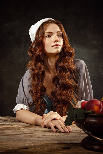 Vintage portrait of young adorable redhead girl in image of medieval person, peasant woman in renaissance style dress isolated on dark background. Comparison of eras, beauty, history, art, creativity. - Фото, изображение
