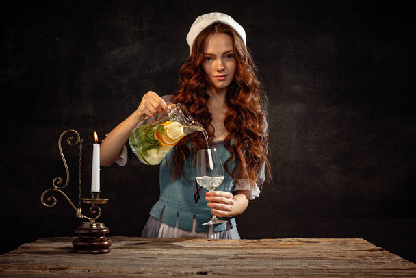 Refreshing drinks. Portrait of young redhead girl with long curly hair in image of medieval person in renaissance style dress isolated on dark background. Comparison of eras, beauty, art, creativity. - Foto, Imagem