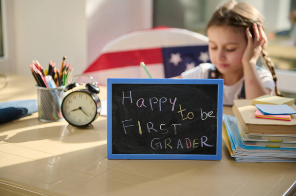 Focus on a chalkboard with text - Happy to be a first grader - against a blurred background of a cute little schoolgirl doing homework while studying remotely from home, in an online school. - 写真・画像