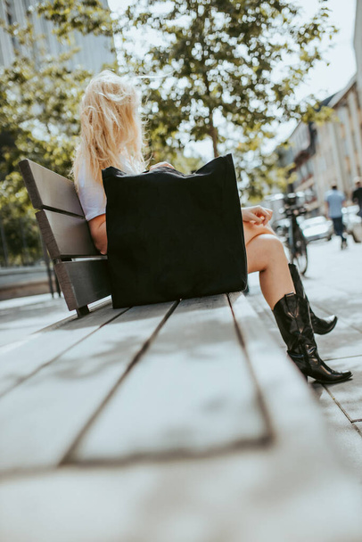 Blonde women sitting on bench at the city with black textile handbag, recycled textile handbag - Foto, Imagen