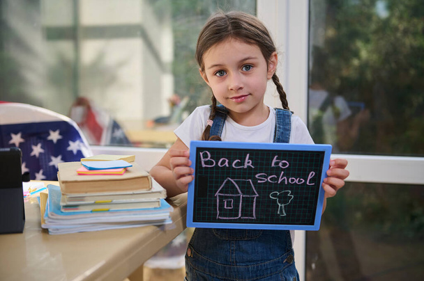 Close-up portrait of an adorable European female kid- schoolgirl in blue denim overalls holding a blackboard with lettering Back to school, standing by a table with folded textbooks and stationary - 写真・画像