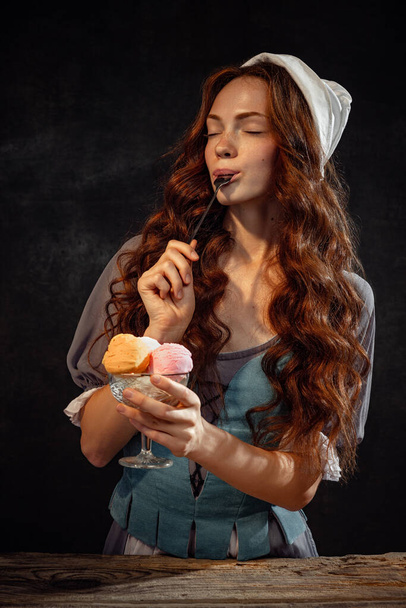 Enjoy the taste. Young beautiful redhead girl with long curly hair in image of medieval person in renaissance style outfit isolated on dark background. Comparison of eras, beauty, art, creativity. - Photo, Image