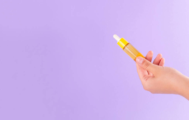 glass yellow bottle with pipette with essential oil in female hand on purple background top view. Aromatic cosmetic product for skin hair care Moisturizing beauty product with vitamin C close up - Foto, Imagen
