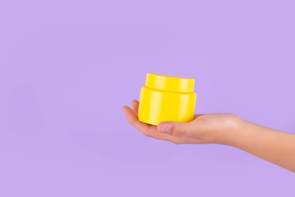 Advertising yellow container, jar in female hand on purple background. Skincare, bodycare beauty product in plastic pot package concept with vitamin c. Yellow unbranded, balsam, face or hand creme jar - Photo, Image
