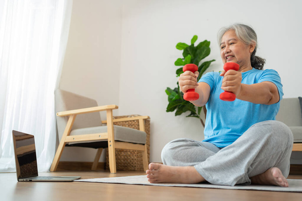 Asian senior woman lifting dumbbell for exercise and workout at home. Active mature woman doing stretching exercise in living room. Exercise Active and healthy for older, elder, and senior concept. - Foto, Bild