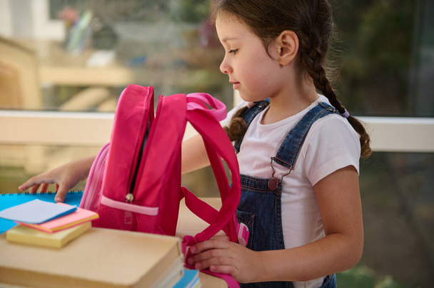 Inspired European first grader, schoolgirl in denim overalls, folding notebooks and school supplies in a pink knapsack, getting ready for the first grade of elementary school, early in the morning - 写真・画像