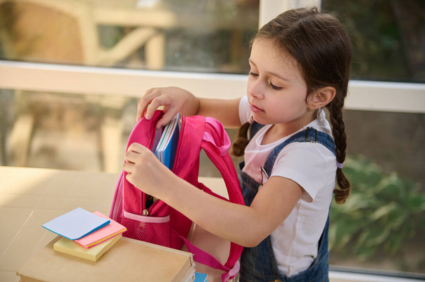 Delightful and inspired schoolgirl in casual denim overalls, puts a notebooks and school supplies in a pink school bag, getting ready for the first grade of elementary school. Back to school concept - Foto, afbeelding