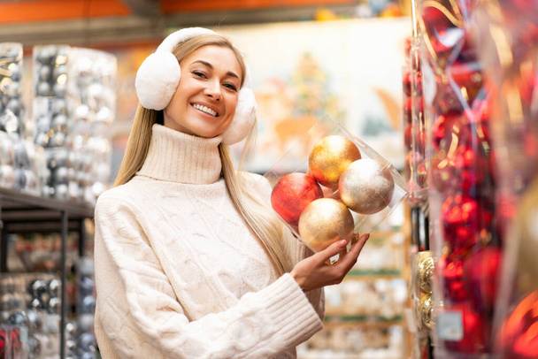 Christmas shopping. Woman choose Christmas decor in supermarket. Caucasian female in fur headphones dressed white sweater buying Christmas ornament. Prepare for winter Holiday. Positive smiling person - Foto, imagen