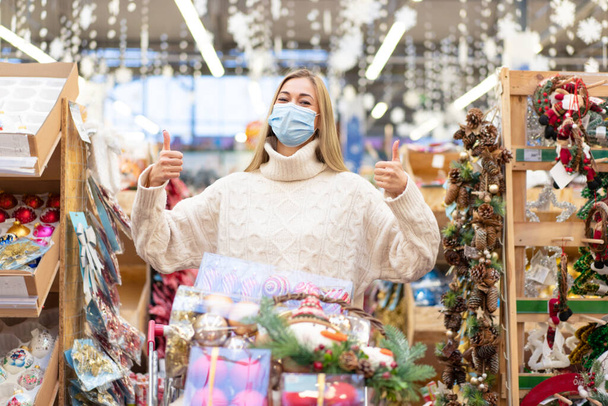 Christmas shopping. Woman with cart shopping in protective medical mask watching Christmas ornament in shopping mall on Holiday sale. Caucasian adult blonde female 30 year choosing Christmas decor - Fotó, kép