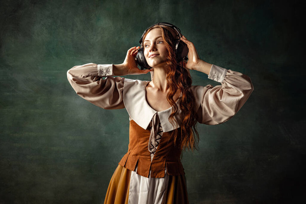 Listening to romantic music. Young beautiful redhair girl with long curly hair like girl of renaissance eras isolated on dark background. Comparison of eras, beauty - Foto, imagen