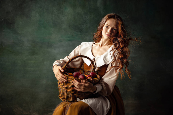 Girl with plums. Young charming redhair girl with long curly hair like girl of renaissance eras isolated on dark background. Comparison of eras, beauty, history, art, creativity. - Foto, afbeelding