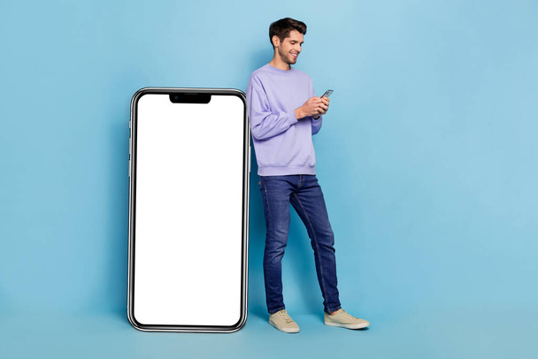 Full size photo of funny millennial brunet guy with telephone wear sweater jeans shoes isolated on blue background. - Foto, Imagen