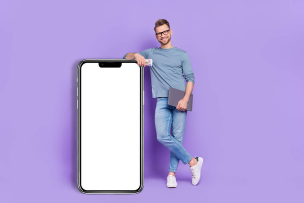Photo of cute handsome guy wear grey shirt holding cash modern device empty space isolated violet color background. - Foto, afbeelding