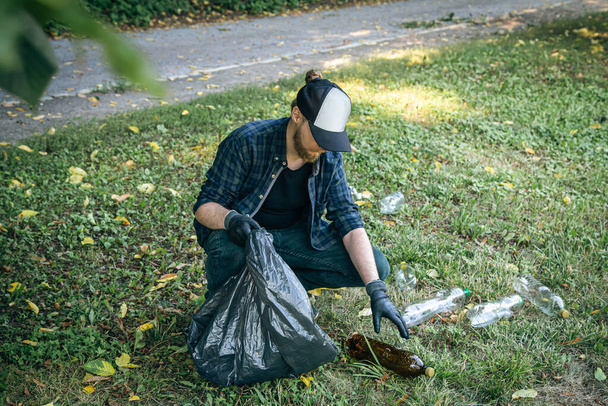 Молодий чоловік зі сміттєвою сумкою в лісі очищає пластикові пляшки, концепцію любові до природи і догляду за екологією
. - Фото, зображення