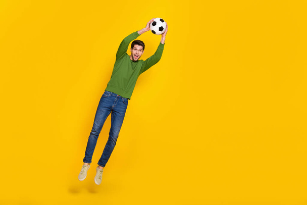 Photo of excited pretty guy wear green sweater jumping high catching ball empty space isolated yellow color background. - Foto, imagen